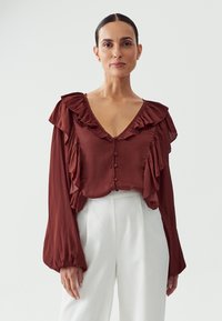 Brown, ruffled blouse with a V-neck and button front. Flowing long sleeves and contrasting white high-waisted trousers. Smooth fabric texture.