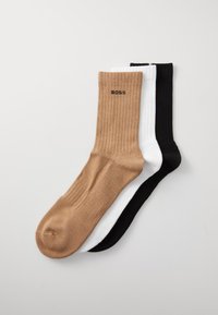 Trois paires de chaussettes côtelées de couleur beige, blanche et noire. Chaque chaussette présente un logo sur le revers et une texture lisse.