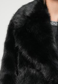 Svart kåpe i kunstpels med myk, luftig tekstur og bred krage. Dekorert med en juvelbesatt pynt nær halsen.