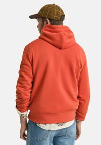 Rode katoenen hoodie met een capuchon en ribgebreide manchetten, gedragen over een bloemenhemd, gecombineerd met lichtblauwe jeans en een geruite pet.