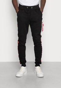 Svarta sweatpants med dragsko i midjan, sidostripes i röd och fickor med dragkedja. Kombineras med vita sneakers. Slät textur.