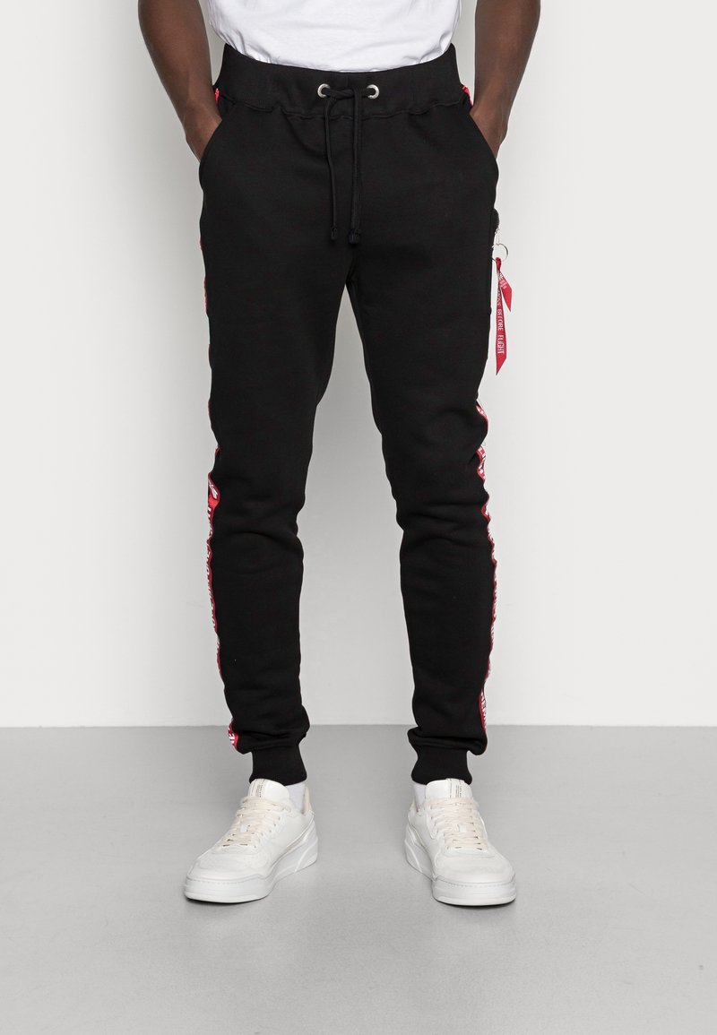 Svarta sweatpants med dragsko i midjan, sidostripes i röd och fickor med dragkedja. Kombineras med vita sneakers. Slät textur.