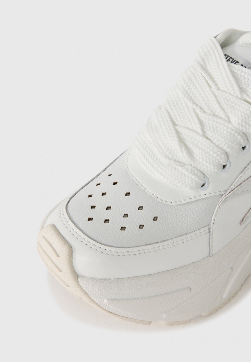 Weißer Sneaker mit diamantförmigen Belüftungslöchern an der Zehenpartie, dicker Sohle und breiten, strukturierten weißen Schnürsenkeln vor einem schlichten Hintergrund.
