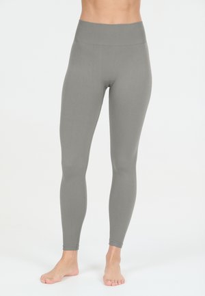 Persoon die hoog getailleerde, effen grijze leggings draagt en blootsvoets staat tegen een effen lichte achtergrond.