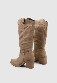 Botas de ante beige hasta la rodilla con un sólido tacón cuadrado, que presentan una cremallera lateral y costuras decorativas en la parte superior.
