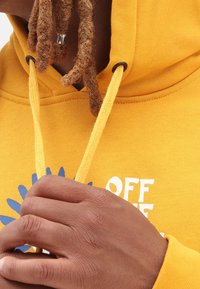 Yellow hoodie with a textured fabric, featuring a front logo in white and blue. Drawstrings in matching yellow, with black metal eyelets.