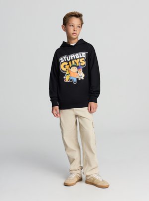 Felpa nera con grafica "STUMBLE GUYS". Pantaloni cargo beige con tasche laterali. Sneakers color cuoio. Il modello posa con un'espressione neutra.
