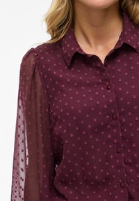 Blouse bourgogne avec un devant boutonné, ornée de pois texturés et de manches transparentes. Design du col et tissu doux et lisse.