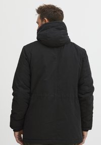 Črna podložena jakna z veliko kapuco, ki ima gladko teksturo in rahlo zoženo obliko, vidne šive ter brez izrazitih vzorcev.