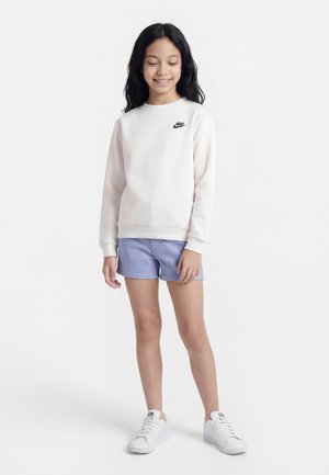 Pige iført en hvid Nike-sweatshirt, lyse lilla shorts og hvide sneakers, stående med lukkede øjne og smilende mod en hvid baggrund.