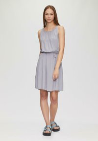 Helles graues ärmelloses Kleid mit leichter Raffung am Ausschnitt und einem taillierten Gürtel. Kombiniert mit blauen Sandalen mit Riemen und einer klobigen schwarzen Sohle.