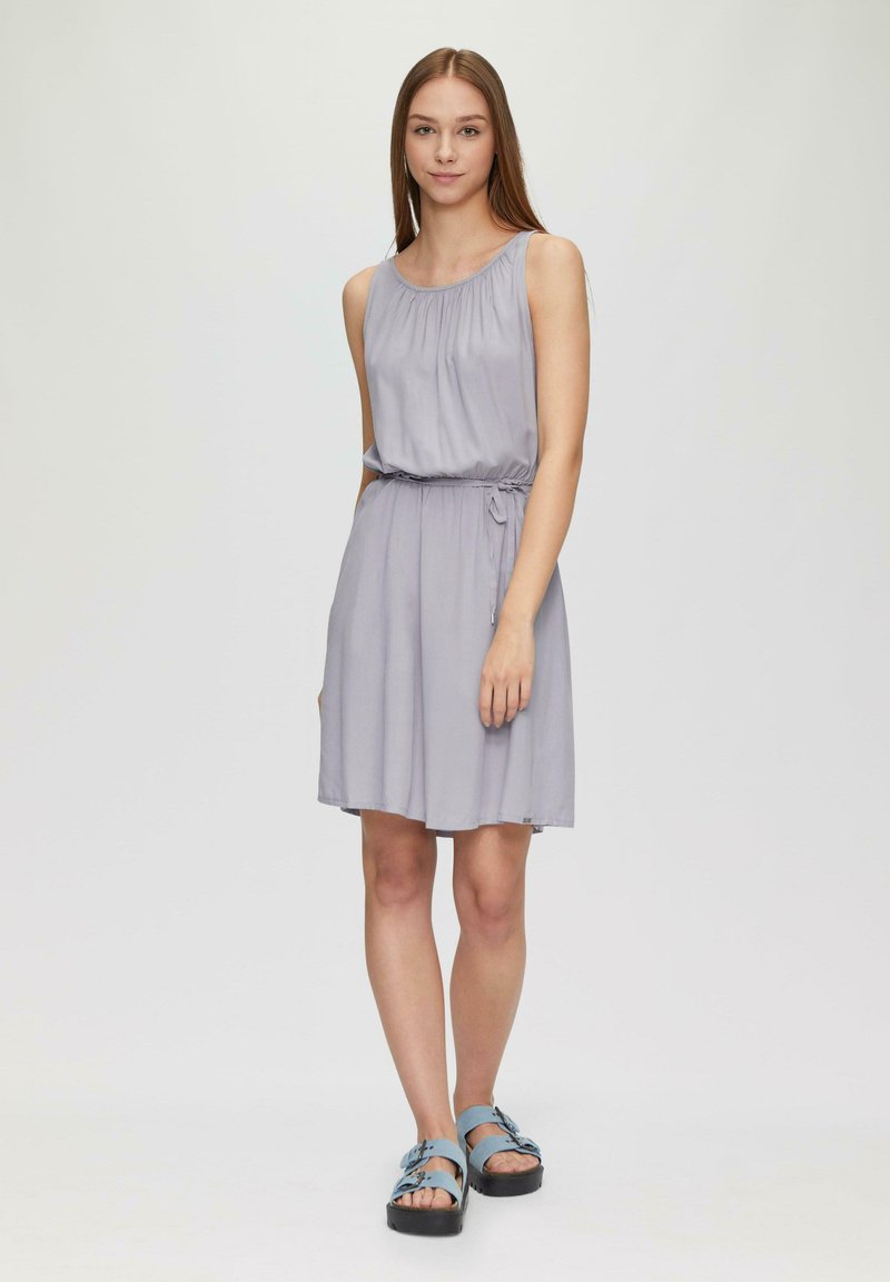 Helles graues ärmelloses Kleid mit leichter Raffung am Ausschnitt und einem taillierten Gürtel. Kombiniert mit blauen Sandalen mit Riemen und einer klobigen schwarzen Sohle.