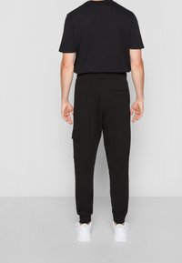 Zwarte sweatpants met een taps toelopende pasvorm, elastische tailleband, zijzakken en een zak op het linkerbeen. Gedragen met een zwart T-shirt en witte sneakers.