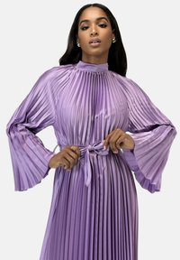 Lilla plisseret maxikjole med høj hals, brede klokkeærmer og bindebælte i taljen. Glat, skinnende tekstur.