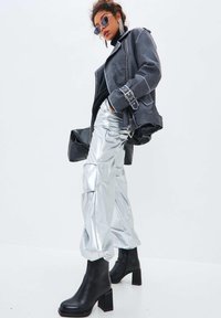 Pantalones plateados y arrugados de gran tamaño, botas de plataforma negras y una chaqueta de cuero gris de motorista con cinturón. Los accesorios incluyen gafas de sol redondas y pendientes grandes.