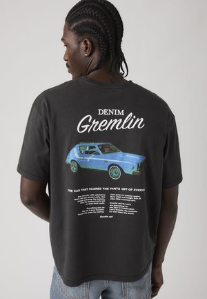 Persona con cabello trenzado viste una camiseta negra que muestra un coche vintage azul y el texto "Denim Gremlin" en la espalda sobre un fondo liso.