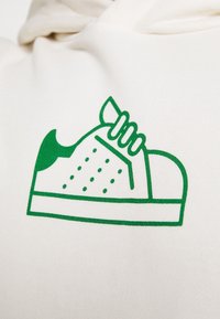 Witte stof met een groene afbeelding van een sneaker met veters, geperforeerde details aan de zijkanten en een stevige basis.