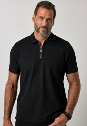 Mand med skæg iført en sort kortærmet polo-shirt med lynlås foran, poserer mod en ensartet lys baggrund.