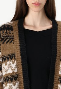 Cárdigan de punto con un patrón geométrico en marrón, blanco y negro. El diseño cuenta con un amplio cuello de canalé negro y un frente abierto.