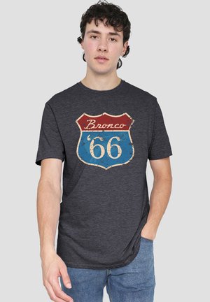 Young man with dark curly hair wearing a dark gray T-shirt with a vintage "Bronco 66" shield logo and blue jeans.