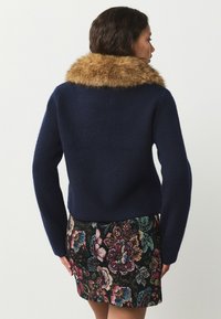 Maglione in lana blu navy con colletto in pelliccia sintetica marrone, maniche lunghe e design aderente, abbinato a una gonna colorata con motivi floreali.