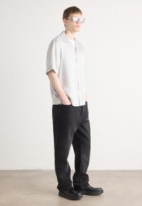 Camicia grigia chiara a maniche corte, con bottoni, abbinata a pantaloni neri larghi e scarpe nere. L'outfit presenta caratteristiche di design minimaliste.