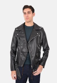 Giacca da motociclista in pelle nera con ampi risvolti, tasche con zip e dettaglio a fibbia laterale. Indossata sopra un maglione a costine color turchese.