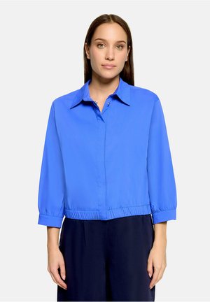 Femme aux cheveux bruns et raides portant une chemise bleu vif à col avec des manches trois-quarts et un pantalon large foncé, debout sur un fond blanc.
