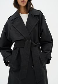 Manteau noir en trench avec un design enroulé, grand col, taille ceinturée et poignets boutonnés, avec une texture de tissu lisse et deux poches.