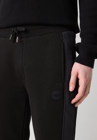 Pantalones jogger negros con cinturilla acanalada y cordón de ajuste, que presentan paneles laterales lisos y un parche de logo circular en el muslo.