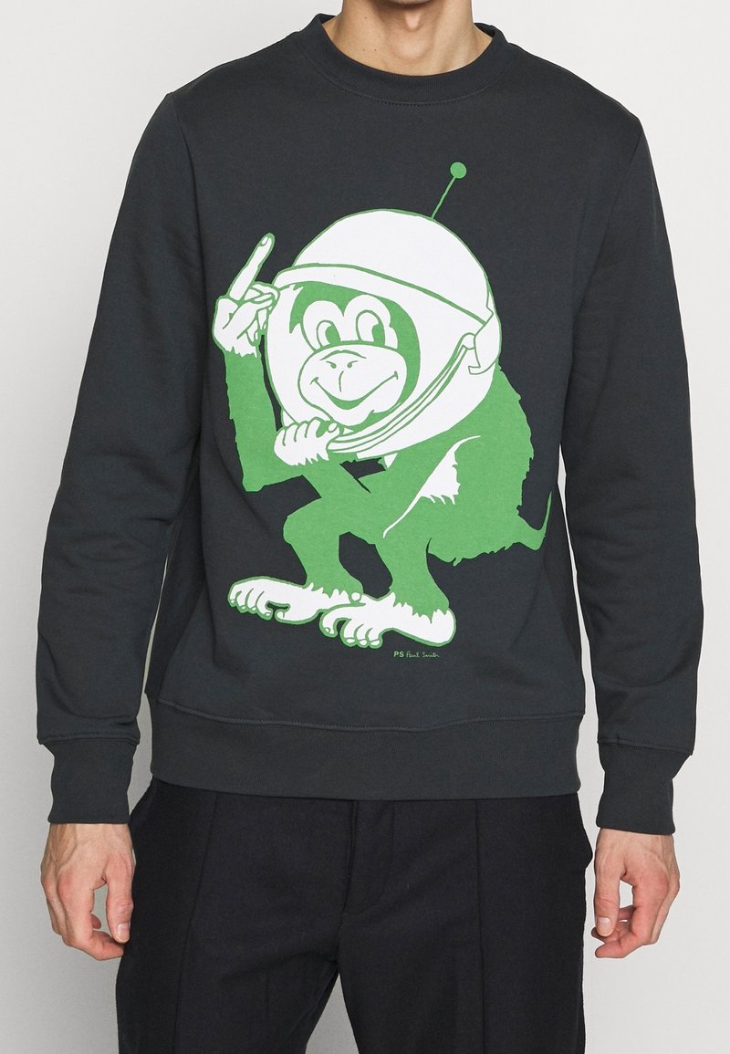 Czarna bluza z dużym zielonym nadrukiem kreskówkowej małpy. Małpa nosi biały hełm i wskazuje palcem, z akcentami na dłoniach i stopach.