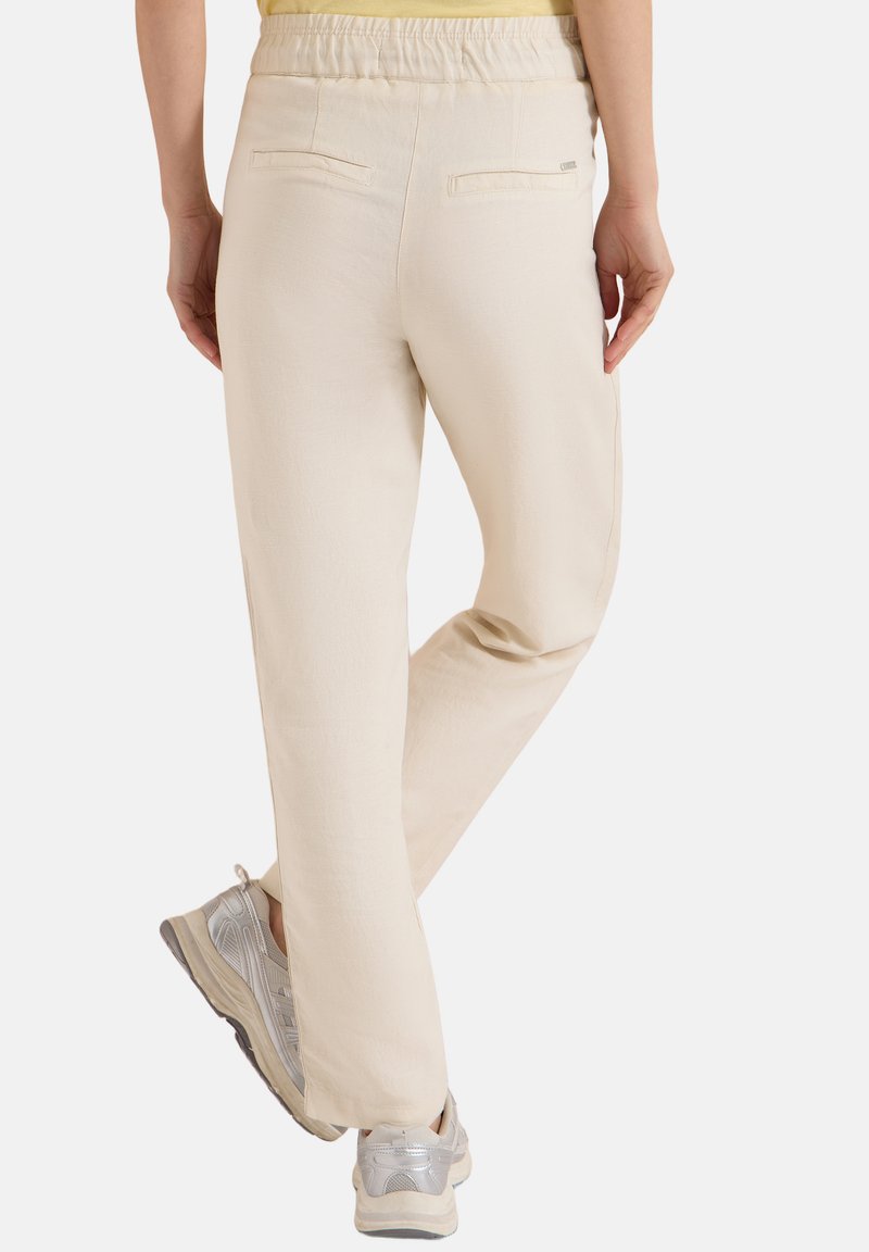 Personne portant un pantalon beige à taille élastique et des baskets argentées, vue de dos avec une jambe légèrement pliée.