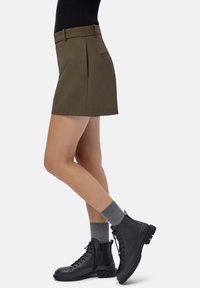 Donna che indossa una mini gonna oliva, calze grigie e stivali neri con lacci alla caviglia, in piedi con una gamba leggermente piegata.