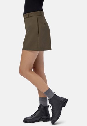 Donna che indossa una mini gonna oliva, calze grigie e stivali neri con lacci alla caviglia, in piedi con una gamba leggermente piegata.