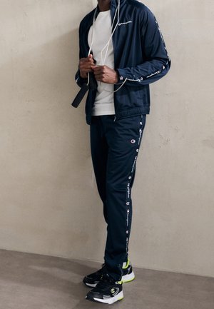 Homme portant un survêtement bleu marine Champion tenant une corde à sauter, debout contre un mur beige uni.