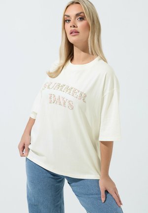 Mujer rubia con camiseta blanca de gran tamaño con texto floral "SUMMER DAYS", combinada con jeans azules, posando frente a un fondo blanco liso.