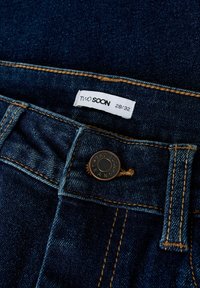 Tamsiai mėlynos džinsinės kelnės turi žalvarinį mygtuką su įrašytu „TWO SOON JEANS“ ir baltą etiketę, rodantį dydį 28/32. Pastebima aukso siuvimo linija.