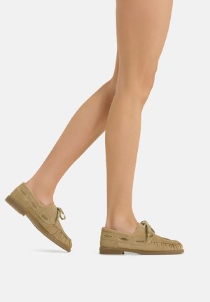 Legs wearing tan suede moccasin-style shoes with laces, photographed in a walking motion against a plain white background.