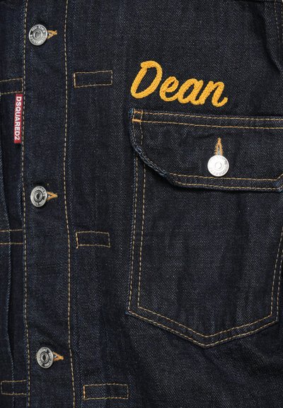 Chemise en denim bleu foncé avec des coutures jaunes, boutons-pression et une poche avant arborant "Dean" brodé en cursive.