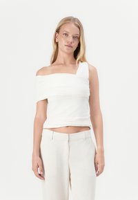 Top blanco de un solo hombro con detalles de tela fruncida, silueta ajustada y textura suave, combinado con pantalones ajustados de color claro.