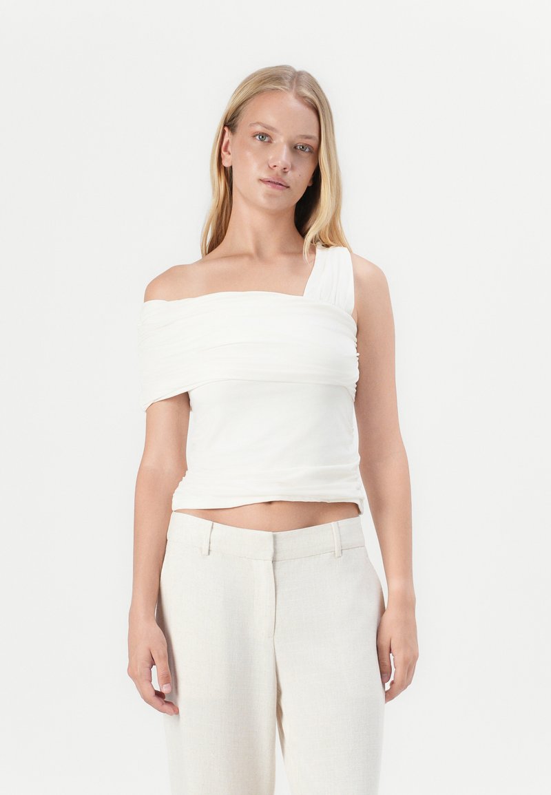 Top blanco de un solo hombro con detalles de tela fruncida, silueta ajustada y textura suave, combinado con pantalones ajustados de color claro.