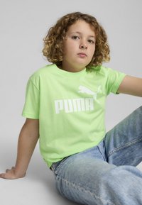 Neona zelena bombažna majica z belim logotipom Puma in napisom. Model jo kombinira s svetlo modrimi jeans hlačami. Tkanina se zdi gladka.