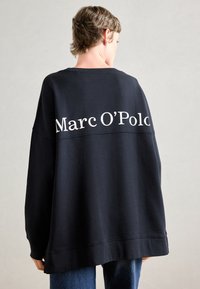 Asmuo su trumpais plaukais, apsisukęs nugarą, vilkintis per didelį juodą džemperį, kuriame viršuje nugaros pusėje baltais rašmenimis parašyta "Marc O’Polo".