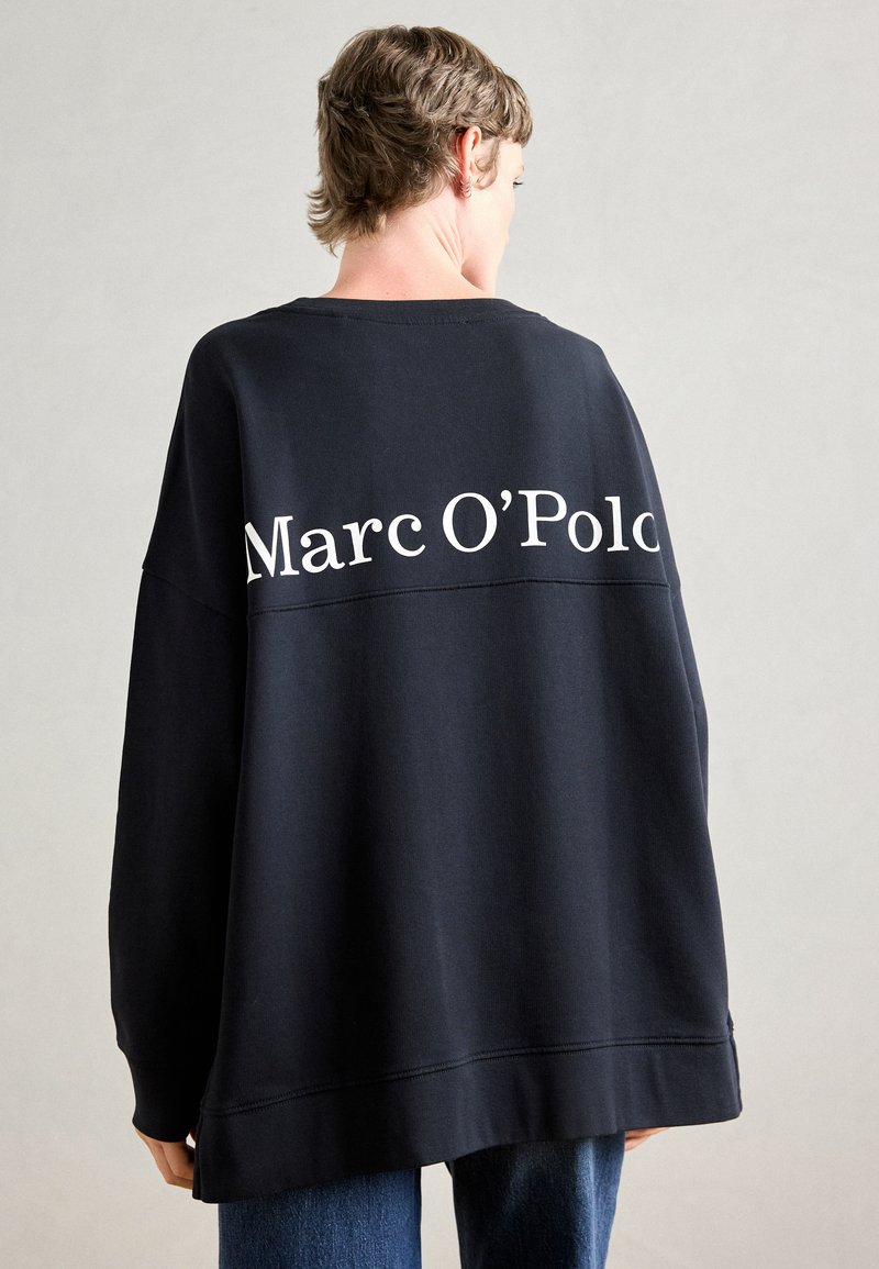 Asmuo su trumpais plaukais, apsisukęs nugarą, vilkintis per didelį juodą džemperį, kuriame viršuje nugaros pusėje baltais rašmenimis parašyta "Marc O’Polo".