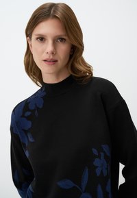 Zwarte trui met een hoge hals en lange mouwen, versierd met blauwe bloemenpatronen aan de linkerkant. Zachte textuur, relaxte pasvorm, minimalistisch ontwerp.