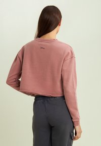 Sudadera de manga larga recortada en tela rosa polvo con puños acanalados. Presenta un logo bordado de "Disney" en la espalda.