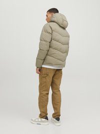 Lys oliven pufferjakke med huva, der har et quiltet design. Kombineret med brune cargo-bukser og hvide sneakers med marineblå detaljer.