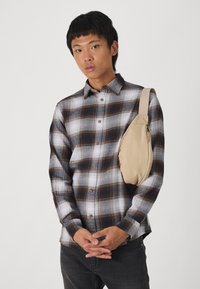 Beige crossoverveske med glatt tekstur, enkelt stropp og glidelås, båret over en svart og hvit rutete skjorte med brune detaljer.
