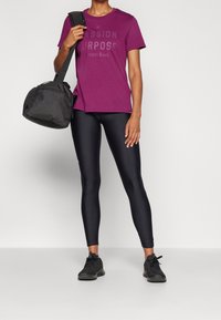 Fialové bavlněné tričko s potiskem "PASSION PURPOSE" v černé barvě, kombinované s černými legínami a černou sportovní taškou. Černé tenisky dotvářejí tento outfit.