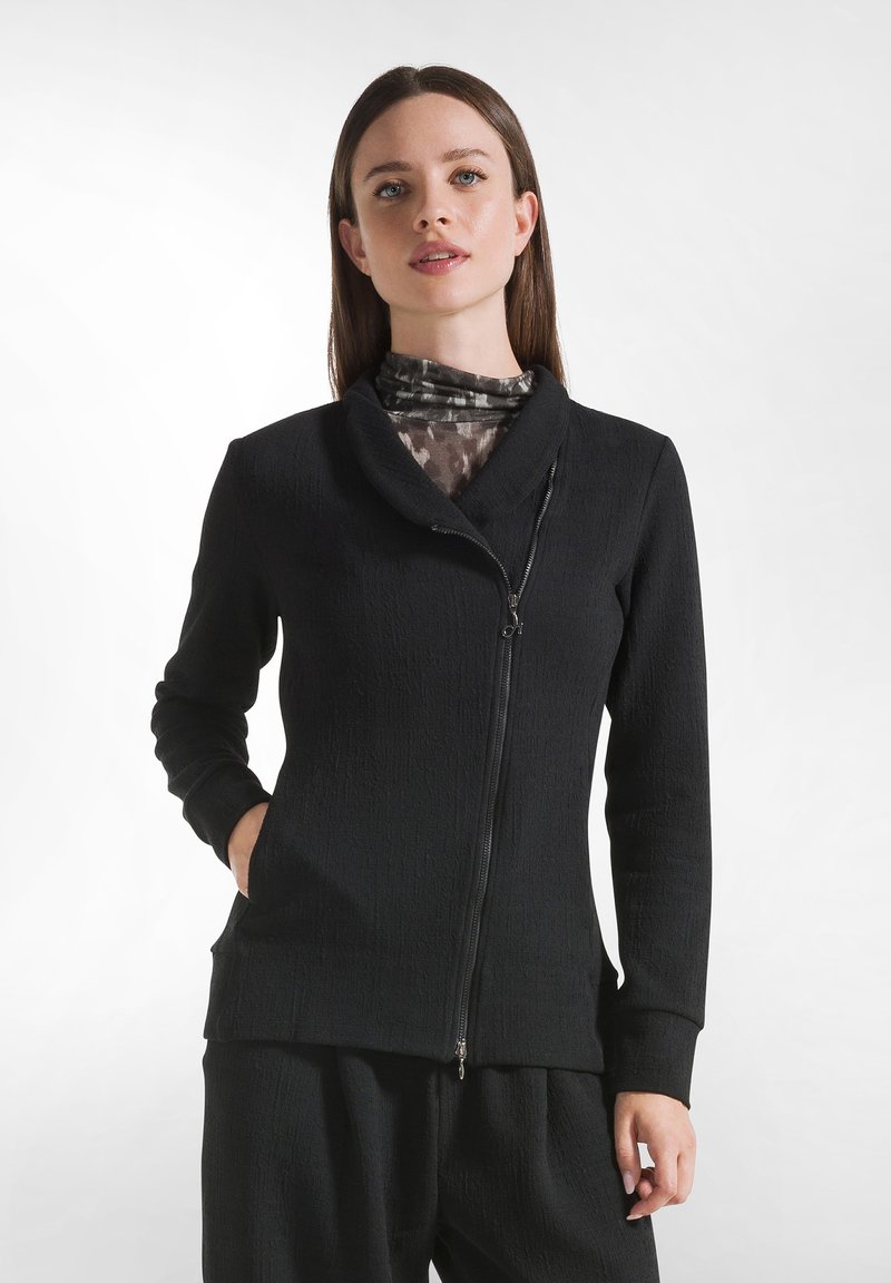 Giacca nera con zip realizzata in un tessuto testurizzato, colletto oversize e due tasche frontali. Indossata sopra un dolcevita a fantasia.